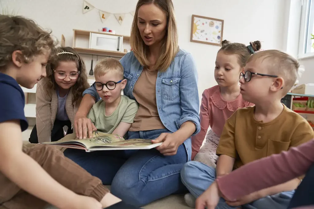A teacher sitting on the floor reading a book with a group of children.